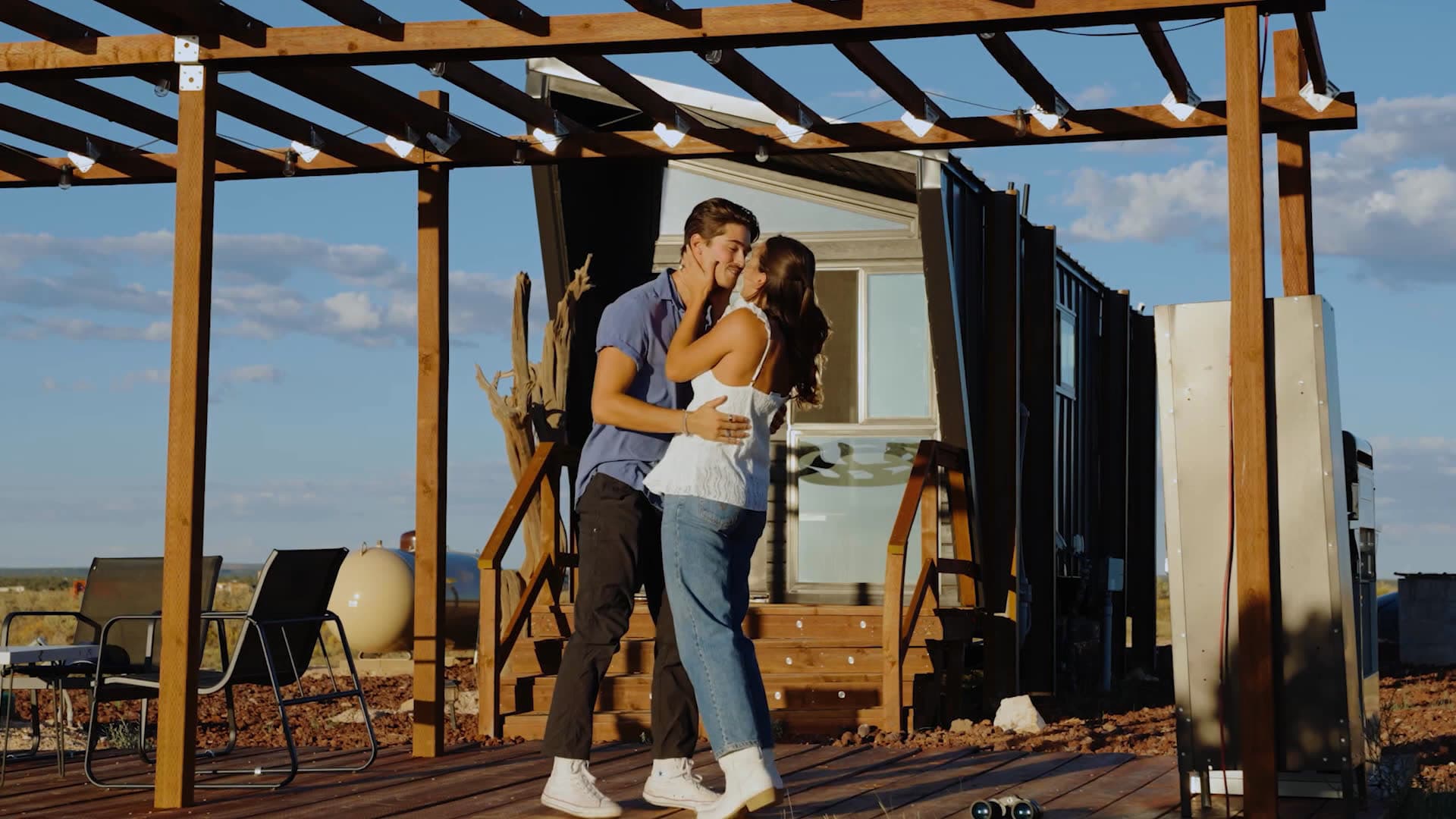 A couple about to kiss under the pergola in front of Jupiter, a dark angular tiny home