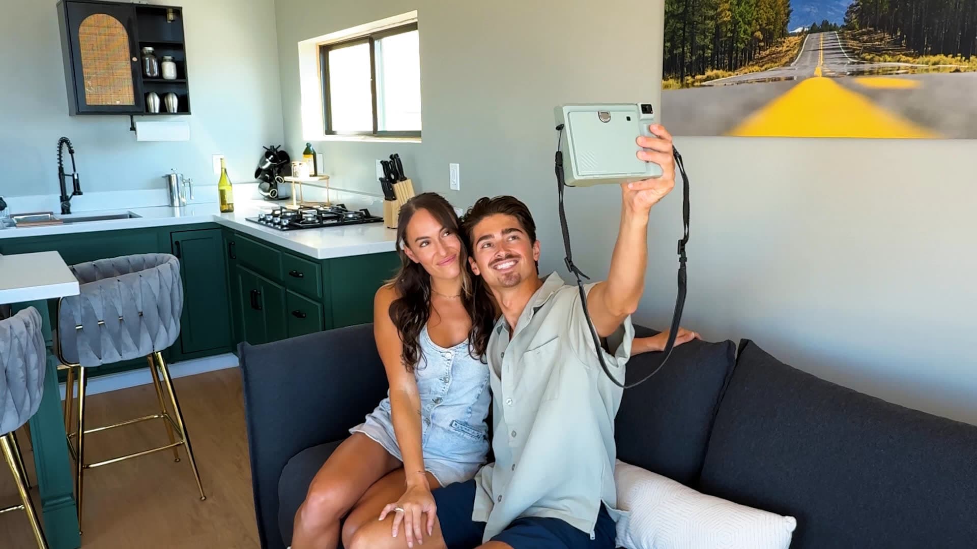 A couple taking a selfie with an instant camera on Luna's sofa, the green kitchen behind them