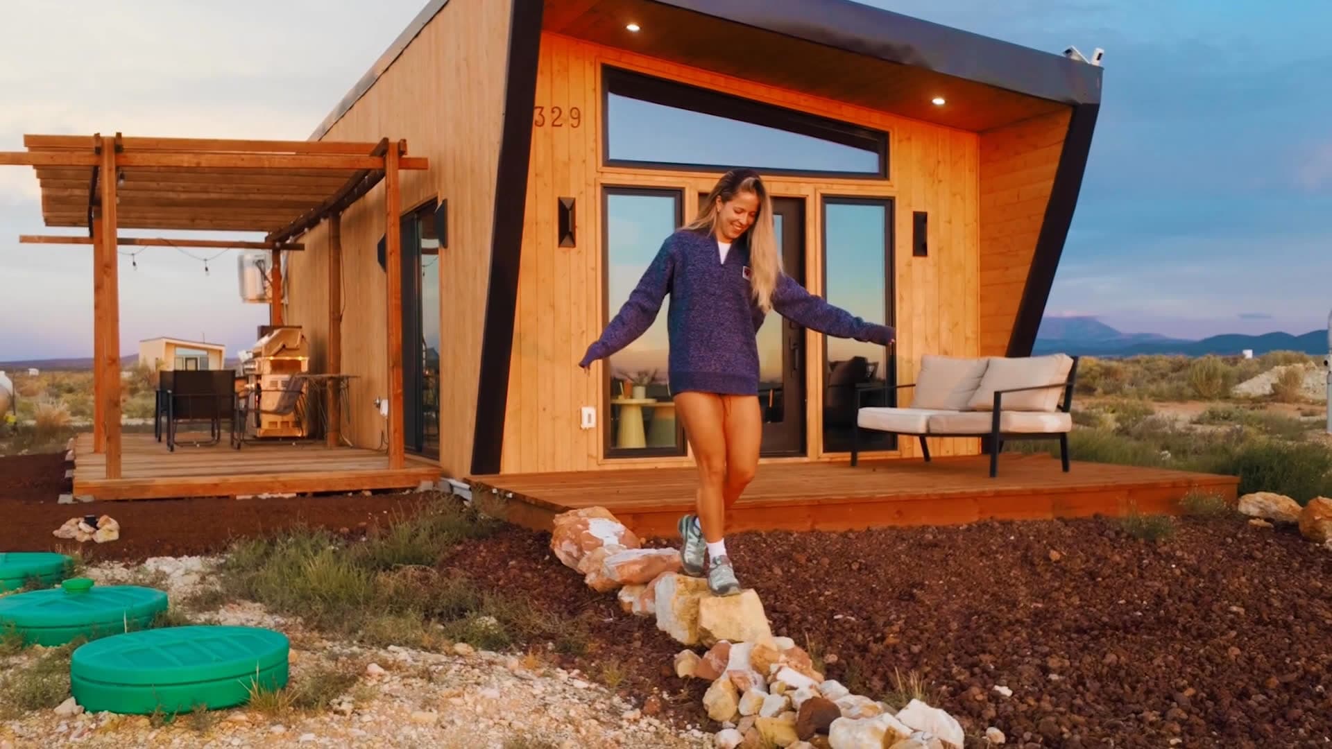 A guest walking along a rock border in front of Venus, a wood-clad tiny home with a pergola deck, at sunset